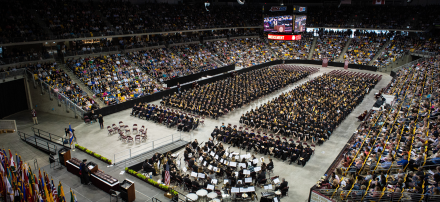 Rows of students in graduation gowns and attendees in an arena sit facing the commencement stage.