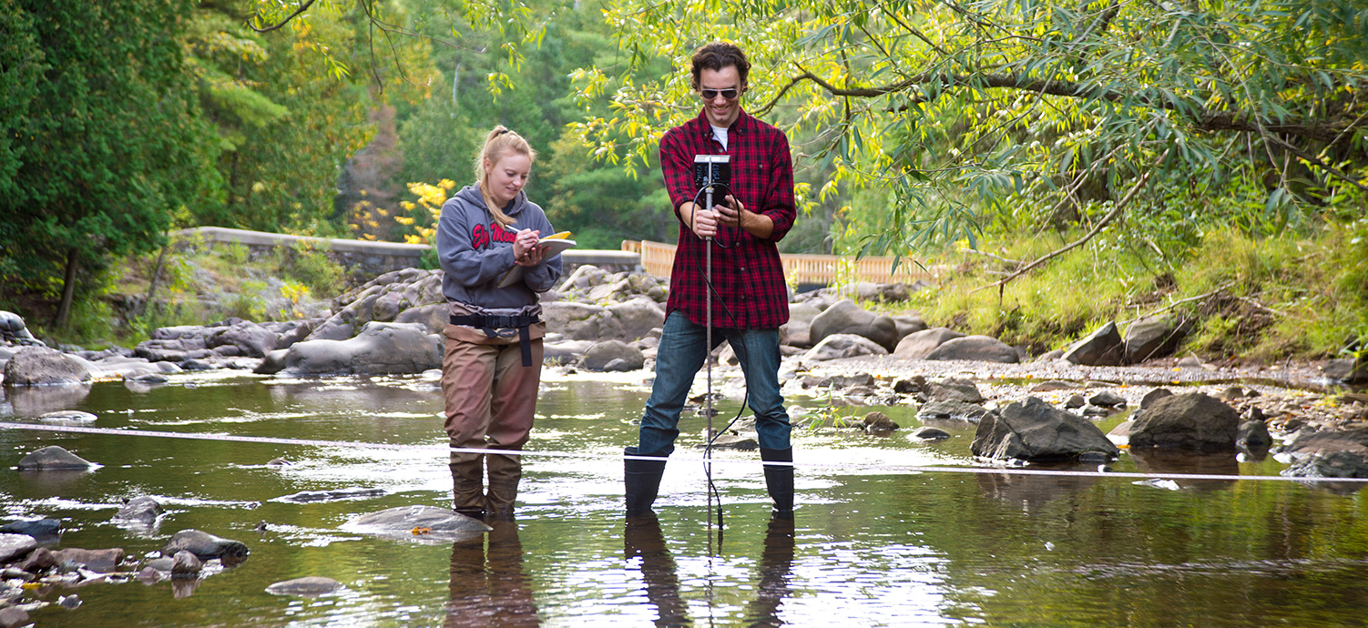 Two students conduct research in a local creek. One student writes on a notepad and the other takes measurements with a tool inserted into the stream.