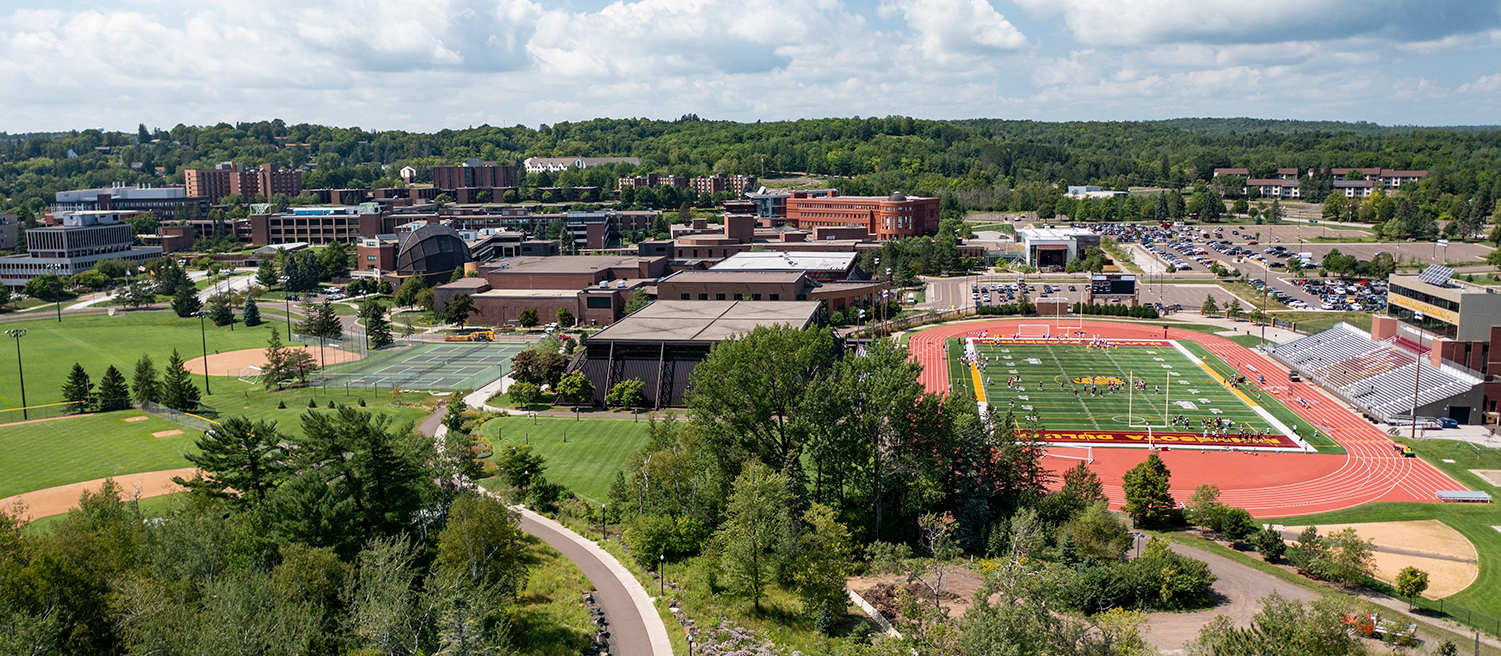 An aerial shot of a campus path winding towards buildings and tennis courts with Malosky Stadium on the right and the wooded hills of Duluth in the background.