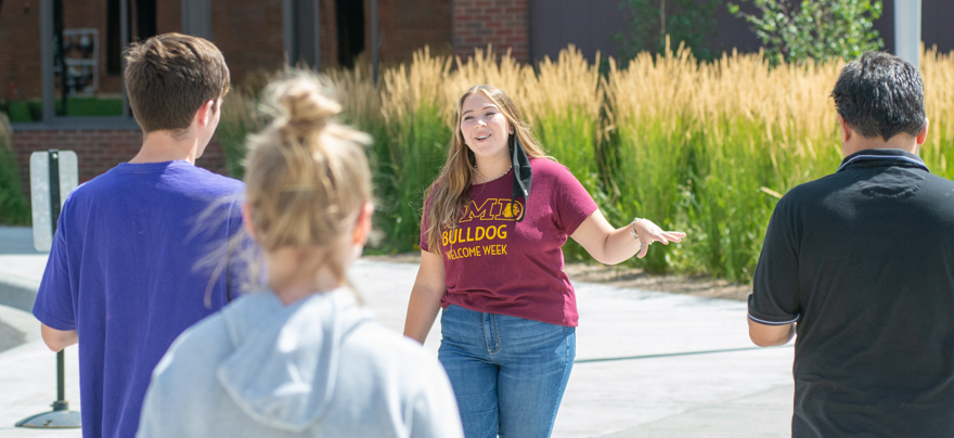 A tour guide motions her arm toward a building on campus as a group of students and parents follows her towards an entrance.
