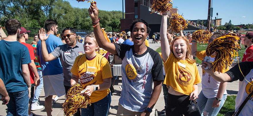Several students cheer in unison as they wave maroon and gold pompoms in the air with smiles on their faces as they welcome incoming students that walk by them out on the football field.